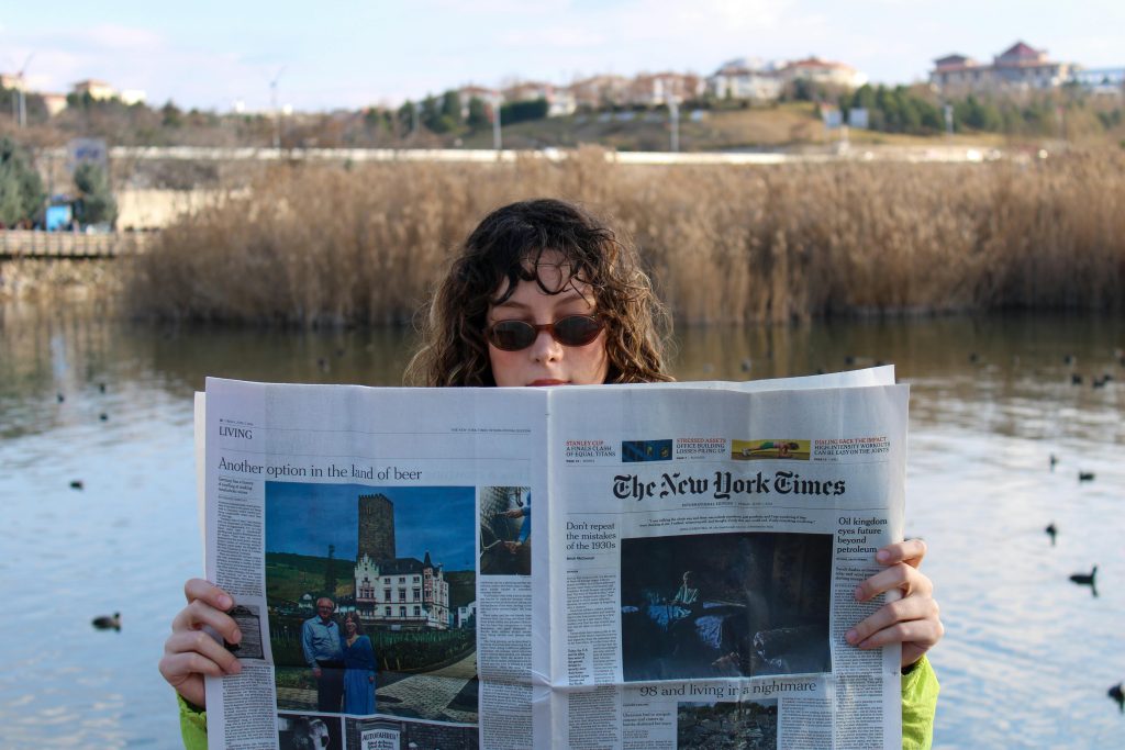 Woman with sunglasses reading a newspaper outdoors by a lake.